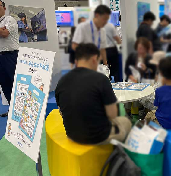 2025年下水道展の明電舎様様のノベルティークリアファイルに描かせて頂いた漫画とイラストです。ノベルティーの看板と来場者の方の様子の写真